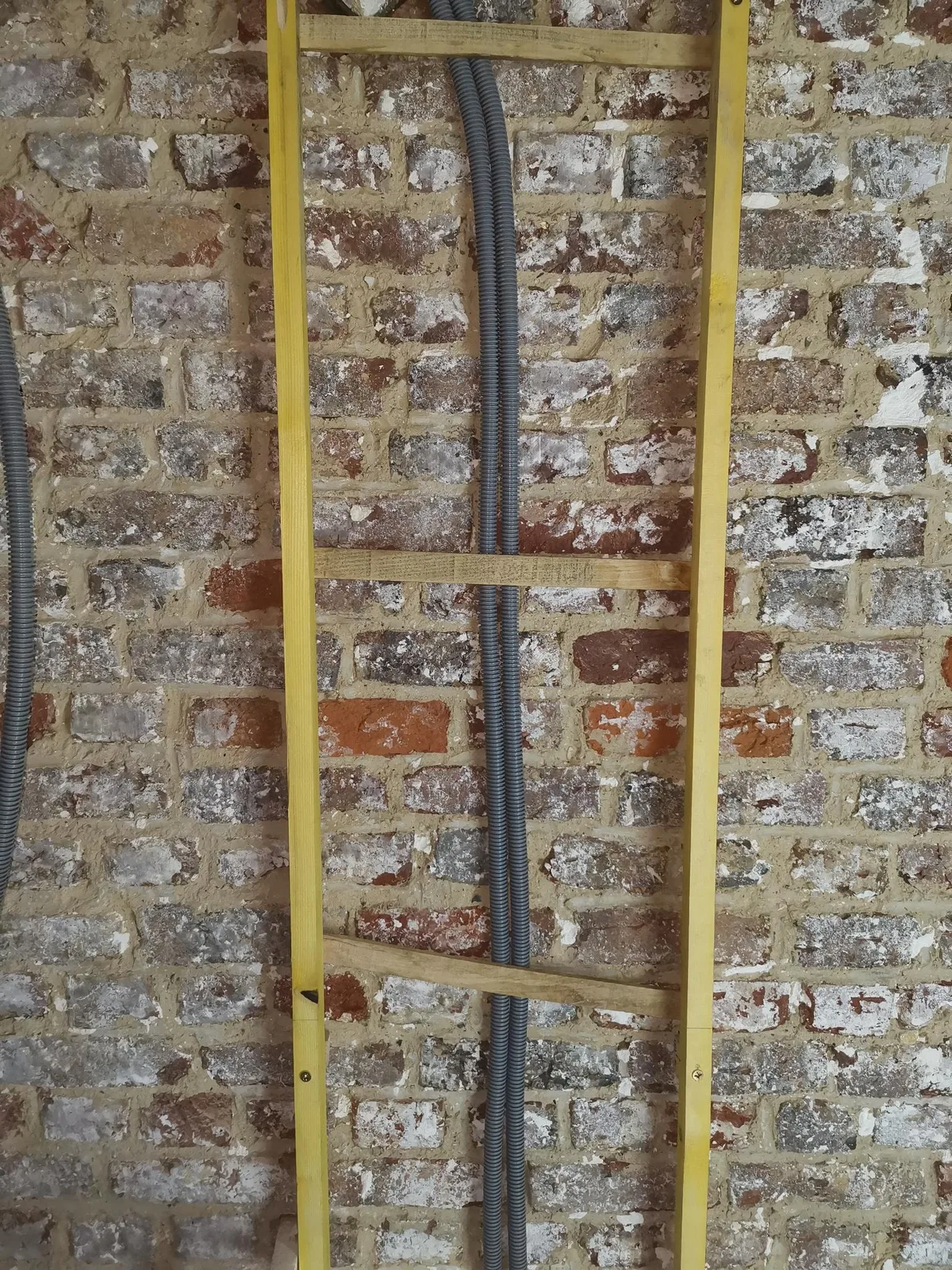Gaine électrique ICTA verticale maintenue contre le mur par de petites cales en bois calées entre deux tasseaux, avant le garnissage en terre-chanvre.