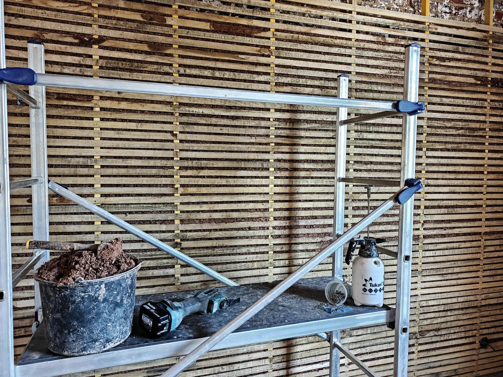 Poste de travail sur l'échafaudage : seau de mélange terre-chanvre, multitool, pulvérisateur à main et serre-joints, devant le mur en lattis.